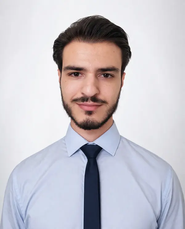 Uomo dal sorriso accennato con capelli e barba scuri che indossa una camicia azzurra e una cravatta blu su uno sfondo bianco