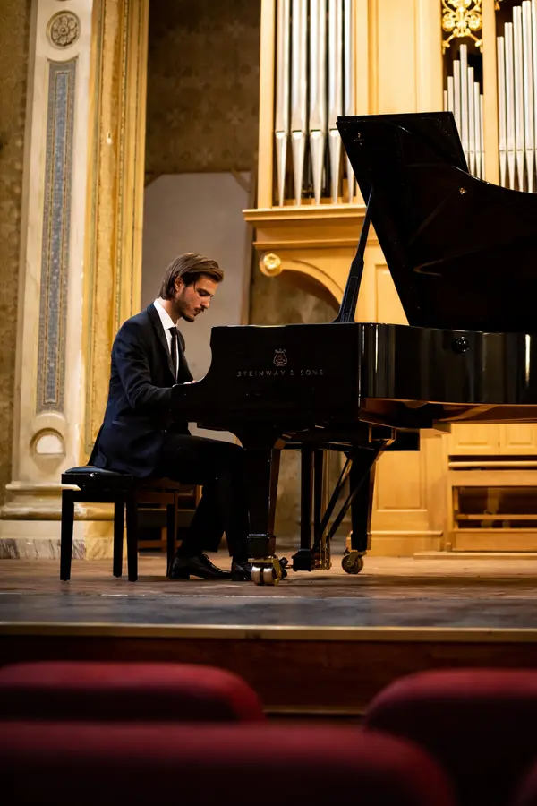 Uomo dal sorriso accennato con capelli e barba scuri che indossa una camicia azzurra e una cravatta blu su uno sfondo bianco